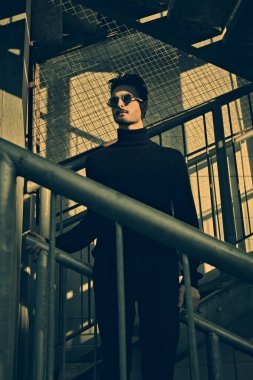Siyah elbiseli ve siyah güneş gözlüklü yakışıklı bir adamın portresi. Metal endüstriyel bir merdivende poz veriyor. Şehirli grunge stili. Erkek modası.