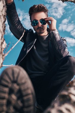 Siyah deri ceketli, güneş gözlüklü, açık havada ağaçta oturan yakışıklı bir genç. Özgürlük ve macera ruhu. Erkek modası. 