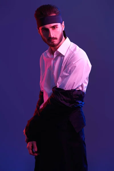Portrait of a handsome masculine young man in black coat, white shirt and with black headband posing in red light. Fighting spirit. Studio shot.