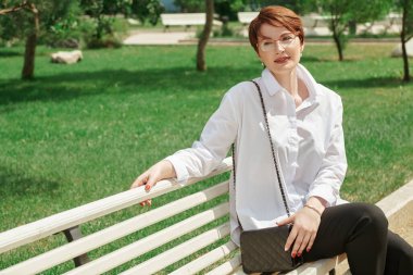 Gözlüklü ve beyaz gömlekli zarif orta yaşlı bir kadın bir yaz günü parkta bir bankta oturuyor. İş kadını portresi.