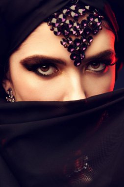 Arap güzelliği, moda. Geleneksel makyaj, siyah başörtüsü ve siyah bir arka planda takıları olan güzel bir doğulu kadının portresi. Makyaj ve kozmetik. 