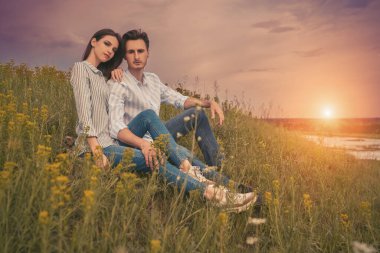 Güzel, romantik bir çift yeşil bir vadide çimlerin üzerinde oturuyor kır çiçekleriyle günbatımının arka planında gökyüzü ve güneş. Aşık insanlar. Yaz zamanı.. 