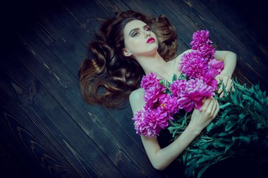 Güzellik ve çiçekler. Parlak makyajlı güzel esmer bir kadının portresi yerdeki şakayık çiçeklerin arasında yatıyor. Makyaj malzemeleri, makyaj malzemeleri. Parfüm.