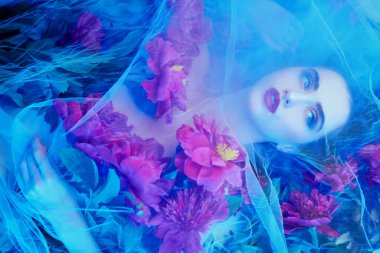 Güzellik ve çiçek konsepti. Parlak makyajlı muhteşem esmer bir kadının portresi. Şakayık çiçeklerinin arasında uzanmış, duvak ile örtülmüş ve sisle çevrili. Makyaj malzemeleri, makyaj malzemeleri. Parfüm.