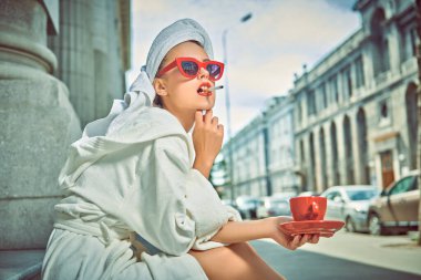 Kafasında beyaz bir havluyla beyaz geceliği ve topuklu ayakkabıları olan çekici bir kadın küçük tüylü köpeğiyle büyük bir şehrin caddesinde yürüyor. Güzellik, moda.