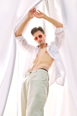 Moda fotoğrafı. Beyaz giysiler ve güneş gözlükleriyle rüzgarda dalgalanan beyaz kumaşlar arasında poz veren yakışıklı genç adam modeli. Yaz stili. Güneş ışığı.