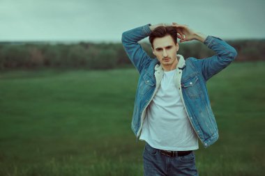 Bulutlu bir günde yeşil vadinin arka planında kot ceketli yakışıklı bir gencin portresi. Erkek güzeli, kot tarzı..