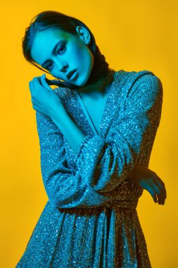 Lüks bir yaşam tarzı. Sarı arka planda parlak yeşil bir ışıkta parlak bir elbiseyle altın makyajla poz veren büyüleyici genç bir kadın. Parti biçimi.