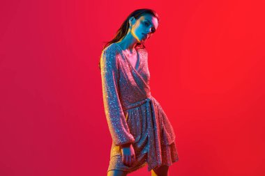 Makyaj ve makyaj malzemeleri. Kırmızı arka planda kırmızı, mavi ve sarı renkli bir ışıkta altın makyajlı muhteşem bir genç kadının portresi. Parti tarzı. Boşluğu kopyala.