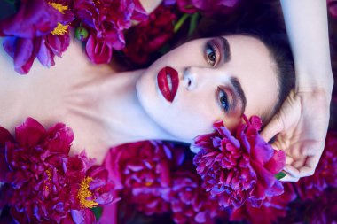 Güzellik ve çiçek konsepti. Şakayık çiçeklerin arasında uzanan parlak makyajlı güzel esmer bir kadının portresi. Makyaj malzemeleri, makyaj malzemeleri. Parfüm.