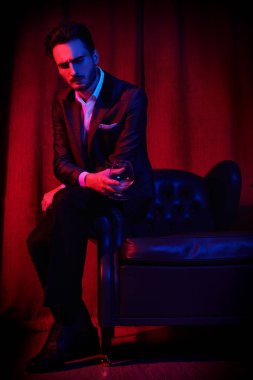 Lüks bir dairede şarap kadehiyle oturan yakışıklı bir esmer adamın portresi. Göz kamaştırıcı bir yaşam tarzı. Moda fotoğrafı. Erkek kıyafetleri ve aksesuarları..