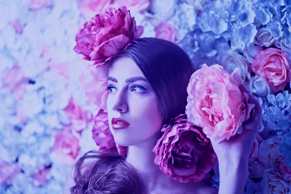 Beautiful young woman with gentle purple lilac make-up and peony flowers in her hair posing on a background of roses. Inspiration of spring and summer. Perfume, cosmetics concept.