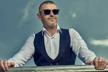 Klasik takım elbiseli ve gökyüzüne karşı modern güneş gözlüğü takmış gülümseyen olgun bir adamın portresi. Erkek tarzı. Erkekler için optik.