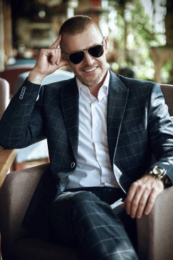 Zarif klasik takım elbiseli yakışıklı olgun bir adam yaz terasında dinleniyor. Erkek modası. Optik, erkekler için güneş gözlüğü. Yaz tatili.