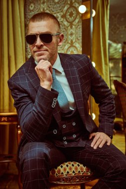 Lüks bir dairede oturan zarif, klasik takım elbiseli, olgun bir adamın portresi. Erkek güzeli, moda. Optik, erkekler için güneş gözlüğü..