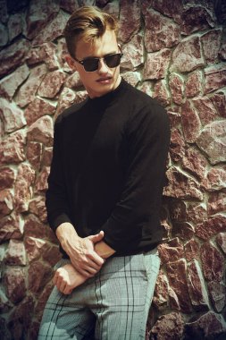 Sanat portresi. Taş duvarın yanında siyah kazak, kot pantolon ve güneş gözlüğü takan yakışıklı genç adam modeli. Erkek stili, güzellik. Moda fotoğrafı.. 