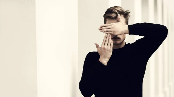 Art portrait. Handsome young man model in black pullover posing next to white columns in the old town, covering his face with his hands. Men\'s style, beauty. Fashion shot.