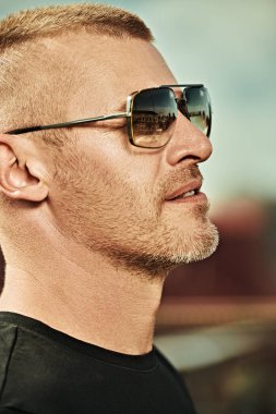 Kısa saçlı ve kıllı yakışıklı, vahşi, olgun bir adamın portresi büyük bir şehrin arka planında moda güneş gözlüğü takıyor. Erkek tarzı. Erkekler için optik.