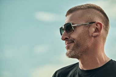 Kısa saçlı ve kıllı yakışıklı, vahşi, olgun bir adamın portresi. Gökyüzünün arka planında güneş gözlüğü takıyor. Erkek tarzı. Erkekler için optik.