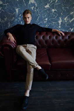 Erkek güzeli, moda. Sarı saçlı yakışıklı bir gencin uzun boylu portresi deri koltukta oturuyor ve hafifçe gülümsüyor..