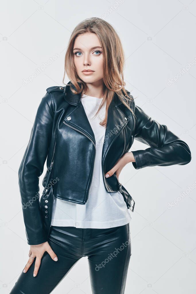 Charming young woman with blonde hair and beautiful blue eyes posing in black leather jacket and leather pants on a white background. Beauty, fashion. 