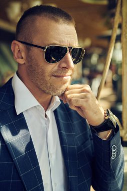 Yakışıklı olgun bir adamın zarif klasik takım elbiseli portresi yaz terasında duruyor. Erkek modası. Optik, erkekler için güneş gözlüğü..