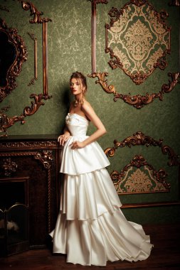 Lüks bir dairede gösterişli beyaz elbiseli ve değerli mücevherlerle poz veren muhteşem bir genç kadın. Klasik klasik iç mimari. Düğün modası.