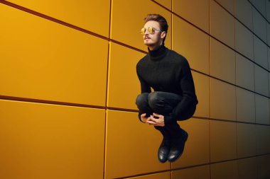 Modern şehir yaşam tarzı. Palto ve güneş gözlüklü şık genç adam sarı sanayi duvarının oradaki bir sokakta koşuyor. Erkek modası. 