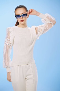 Zarif beyaz elbiseler ve açık mavi arka planda güneş gözlüğü takan güzel bir yaz kızı. Mutlu gülümseyen kız. Yaz modası ve makyaj.