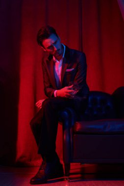 Lüks bir dairede şarap kadehiyle oturan yakışıklı bir esmer adamın portresi. Göz kamaştırıcı bir yaşam tarzı. Moda fotoğrafı. Erkek kıyafetleri ve aksesuarları..