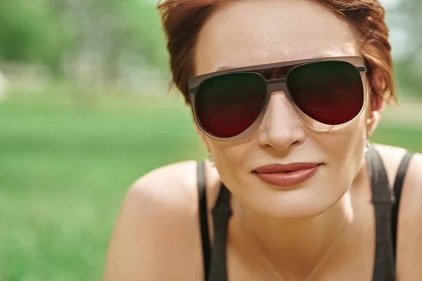 Happy middle-aged woman in modern sunglasses in summer park. Close up portrait.