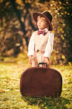Güzel bir sonbahar parkında büyük kahverengi bir bavulla klasik takım elbiseli ve şapka giyen mutlu bir çocuk. Retro tarzı. Çocuk modası.