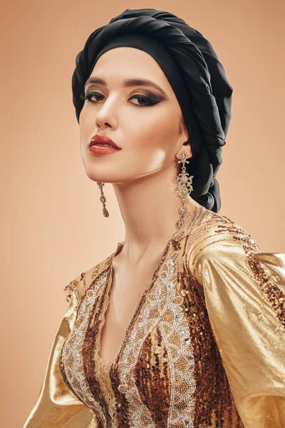 Oriental beauty woman. Portrait of a beautiful arabian woman in golsen top with traditional oriental make-up and black turban. Make-up and cosmetics.