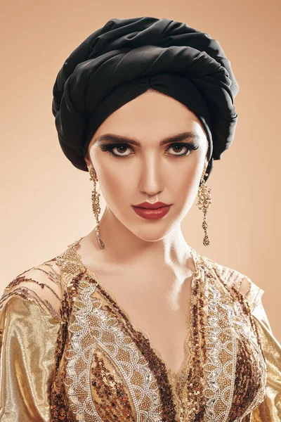 Oriental beauty woman. Portrait of a beautiful arabian woman in golsen top with traditional oriental make-up and black turban. Make-up and cosmetics.