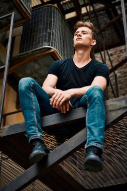 Sıradan giyinmiş yakışıklı genç adam metal endüstriyel bir merdivende oturuyor. Şehirli grunge stili. Erkek modası.