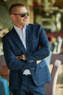 Zarif klasik takım elbiseli yakışıklı olgun adam yaz terasında duruyor. Erkek modası. Optik, erkekler için güneş gözlüğü. Yaz tatili.
