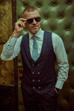 Lüks bir dairede duran şık takım elbiseli, olgun, yakışıklı bir adamın portresi. Erkek güzeli, moda. Optik, erkekler için güneş gözlüğü..