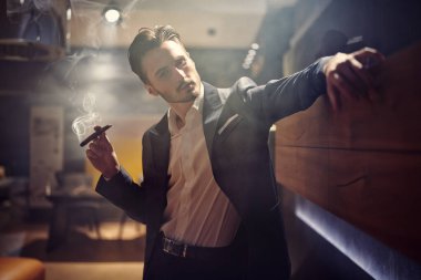 Zarif takım elbiseli yakışıklı esmer adam lüks bir dairede duruyor ve puro içiyor. - Mafya. Erkek güzelliği, moda.