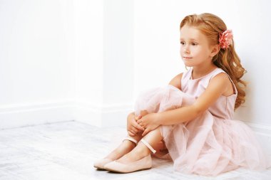 Beyaz bir odada, yerde oturan, güzel, açık renk elbiseli küçük bir kızın portresi. Stüdyo çekimi. Mutlu bir çocukluk. Çocuk modası. 