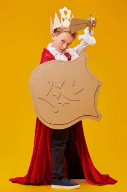 Kartondan zırhı olan, şövalye kostümü giymiş yakışıklı cesur bir çocuğun tam boy portresi. Sarı arka plan. Çocukluk hayalleri ve fantazileri. Karnaval, Tiyatro.