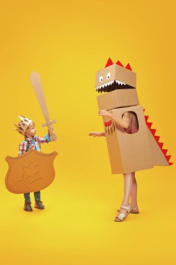 Portrait of a cute little boy in a knight costume with cardboard armour playing with a girl in a dragon costume. Childhood dreams. Full length portrait on a yellow background. clipart