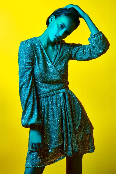 Lüks bir yaşam tarzı. Sarı arka planda parlak yeşil bir ışıkta parlak bir elbiseyle altın makyajla poz veren büyüleyici genç bir kadın. Parti biçimi.