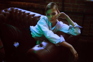 Modern genç kadın şık siyah kadife bir takım elbise ve deri bir kanepede beyaz gömlekle poz veriyor. Lüks bir yaşam tarzı. Moda, makyaj ve güzellik trendleri.