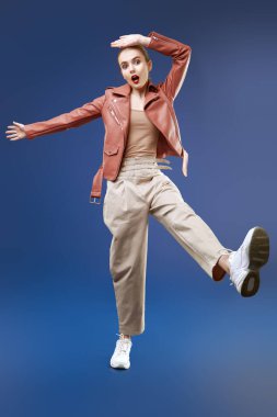 Heyecanlı, pantolonlu, deri ceketli, geniş bir adım atıp koyu mavi arka planda ağzını açan modern bir kız modelin portresi. Güzellik, moda. Boşluğu kopyala. 