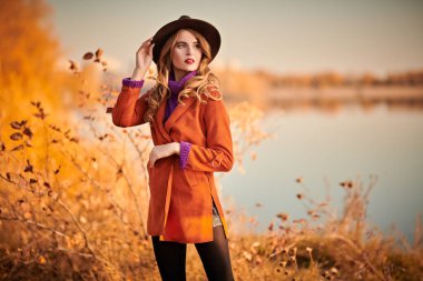 Güz parkında nehir kenarında poz veren güzel bir genç kadın. Sonbahar güzelliği, moda.