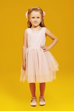 Kıvırcık sarı saçlı ve sarı arka planda poz veren güzel açık renk elbisesi olan tatlı küçük bir kız. Stüdyo çekimi. Çocuk modası. 