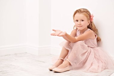 Beyaz bir odada, yerde oturan, güzel, açık renk elbiseli küçük bir kızın portresi. Stüdyo çekimi. Mutlu bir çocukluk. Çocuk modası. 