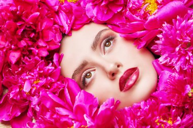 Güzellik ve çiçekler. Şakayık çiçeklerin arasında uzanan parlak makyajlı güzel esmer bir kadının yakın plan portresi. Makyaj malzemeleri, makyaj malzemeleri. Parfüm.