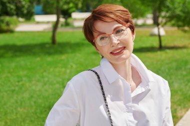 Gözlüklü ve beyaz gömlekli zarif orta yaşlı kadın bir yaz günü parkta poz veriyor. İş kadını portresi. Optik biçimi.