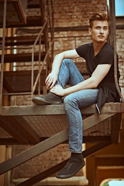 Sıradan giyinmiş yakışıklı genç adam metal endüstriyel bir merdivende oturuyor. Şehirli grunge stili. Erkek modası.